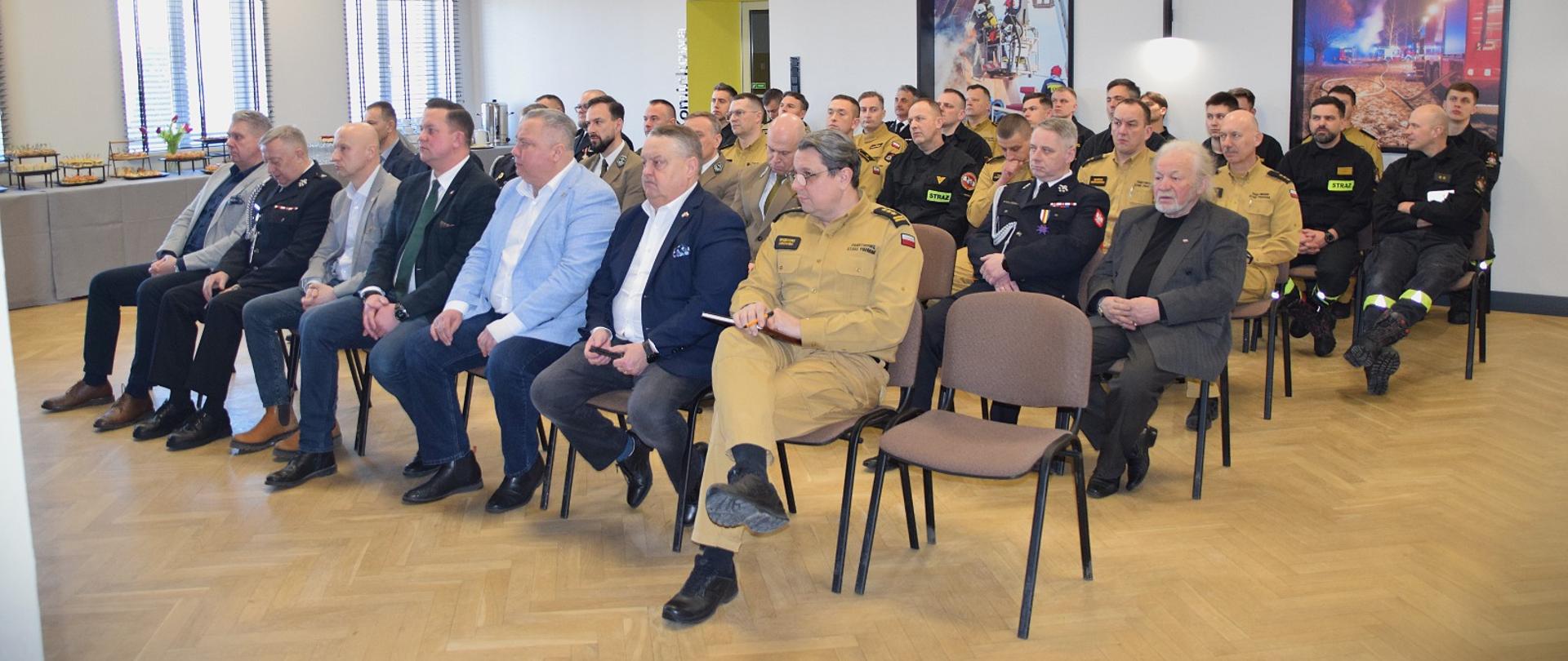 Narada podsumowująca funkcjonowanie ochrony przeciwpożarowej w powiecie monieckim w roku 2025 - zaproszeni goście podczas odprawy siedzą na krzesłach na sali, w tle 2 zdjęcia strażackie w dużych ramach