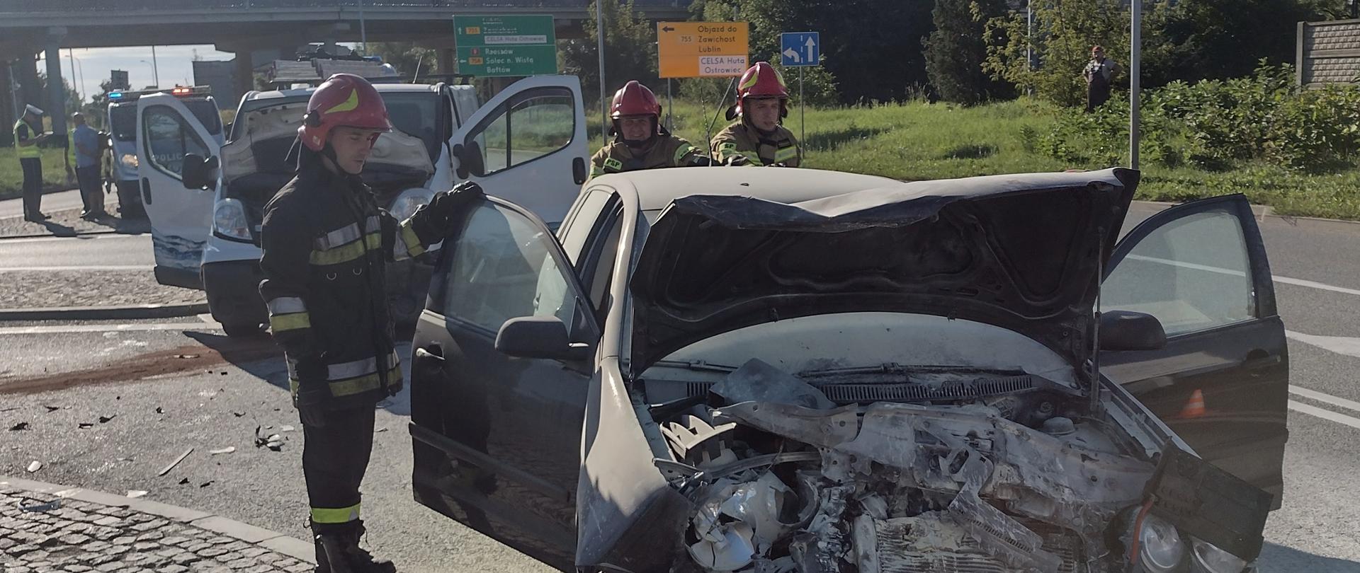 Na pasie jezdni znajduje się uszkodzony samochód osobowy VW Polo. Spalona komora silnika wypełniona jest proszkiem gaśniczym. Obok pojazdu znajdują się 3 strażacy którzy spychają auto na pobocze. W tle drugie uszkodzone auto oraz radiowóz Policji. 