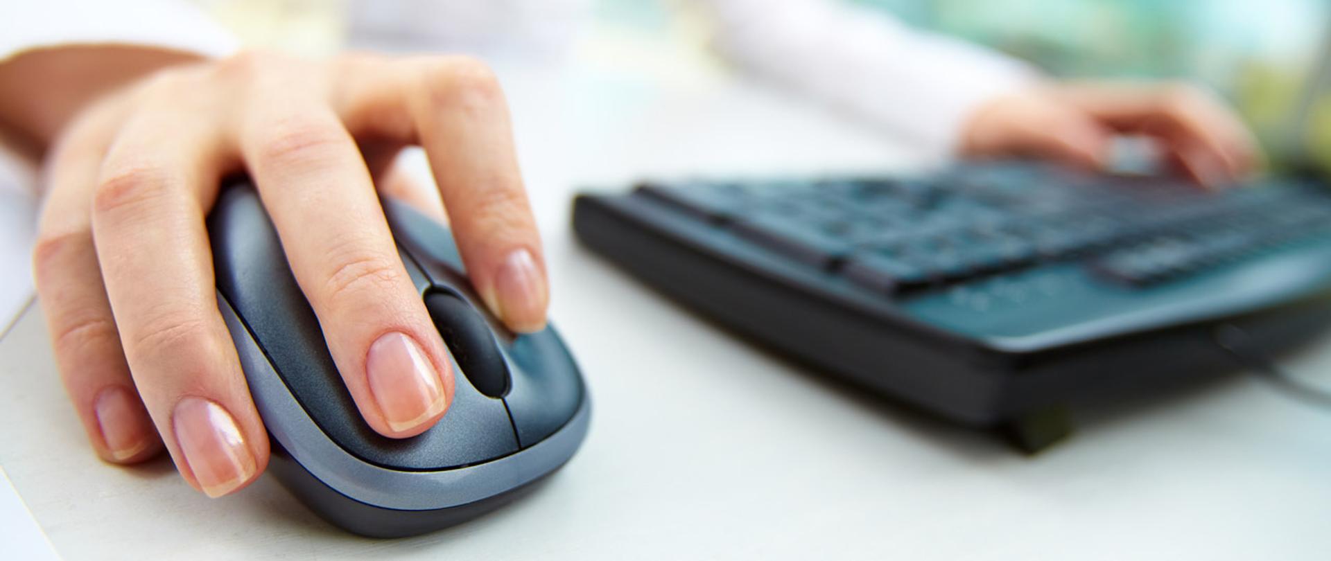 Dłoń na myszy komputerowe, druga na klawiaturze komputerowej.