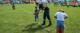Na zdjęciu widać dwoje dzieci oraz strażaka podczas obsługi hydronetki strażackiej. Jedno dziecko pompuje wodę, drugie trzyma prądownicę, strażak asekuruje dziecko. W tle widać piknik rodzinny.