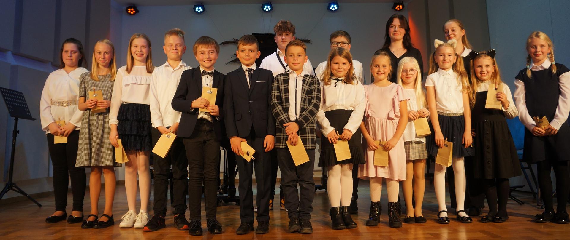 Zdjęcie przedastawia uczniów klas pierwszych stojącycj na scenie szkoły, z upominkami po pasowaniu na ucznia.