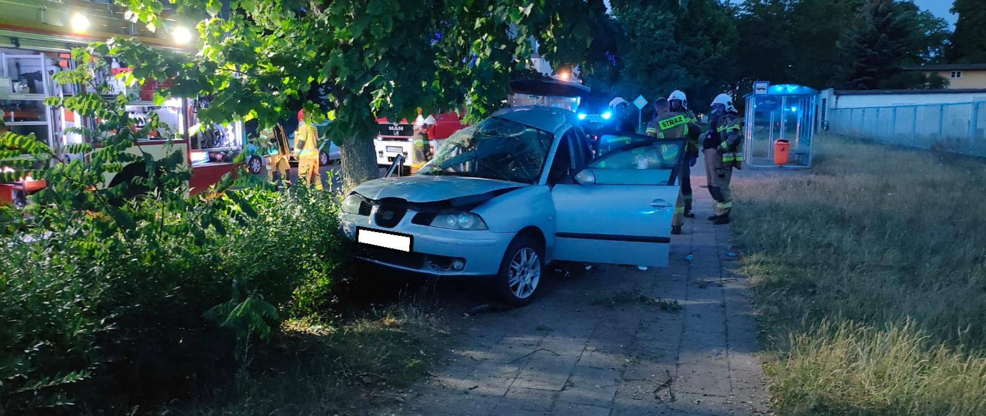Samochód osobowy uderzył w drzewo bokiem i stoi na chodniku.