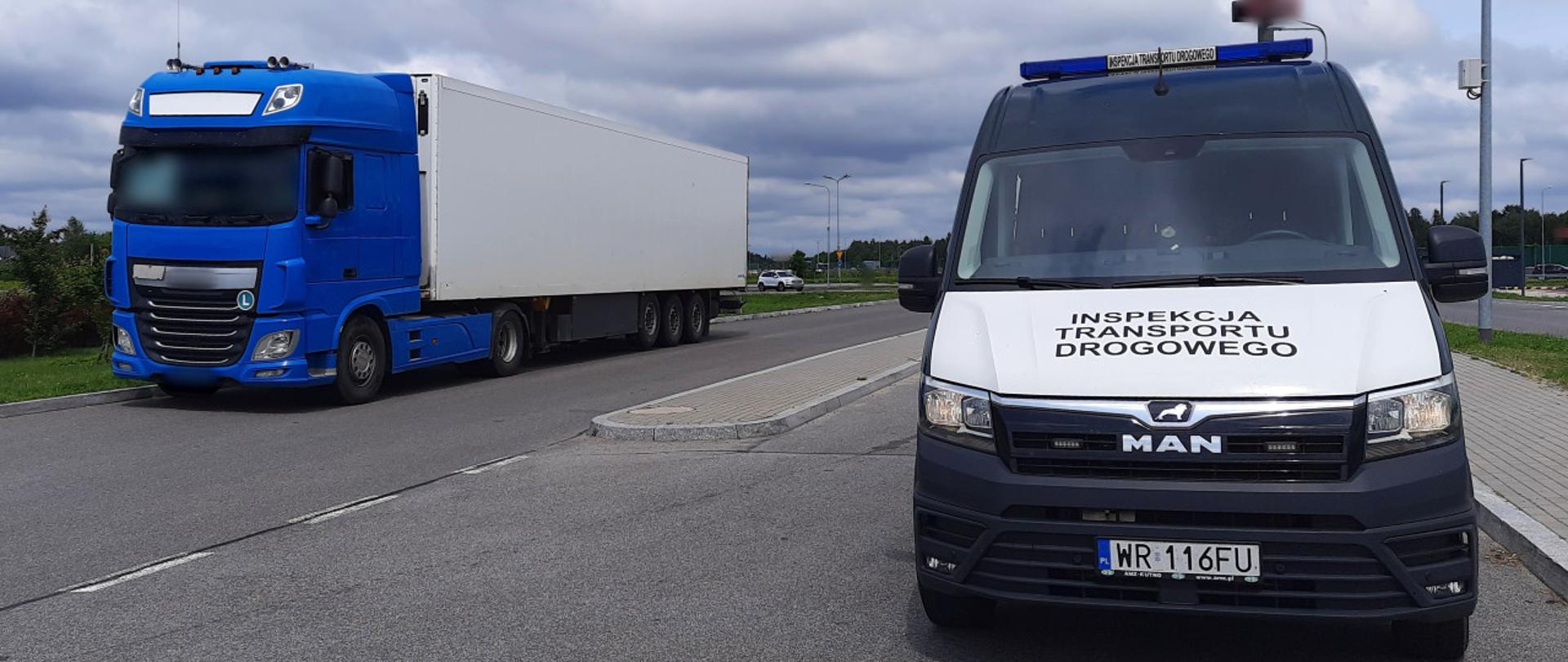 Na pierwszym planie: przód oznakowanego furgonu mazowieckiej Inspekcji Transportu Drogowego. W tle: ciągnik siodłowy z podpiętą naczepą stoi w zatoce kontrolnej, w pobliżu oznakowanego furgonu Inspekcji Transportu Drogowego.