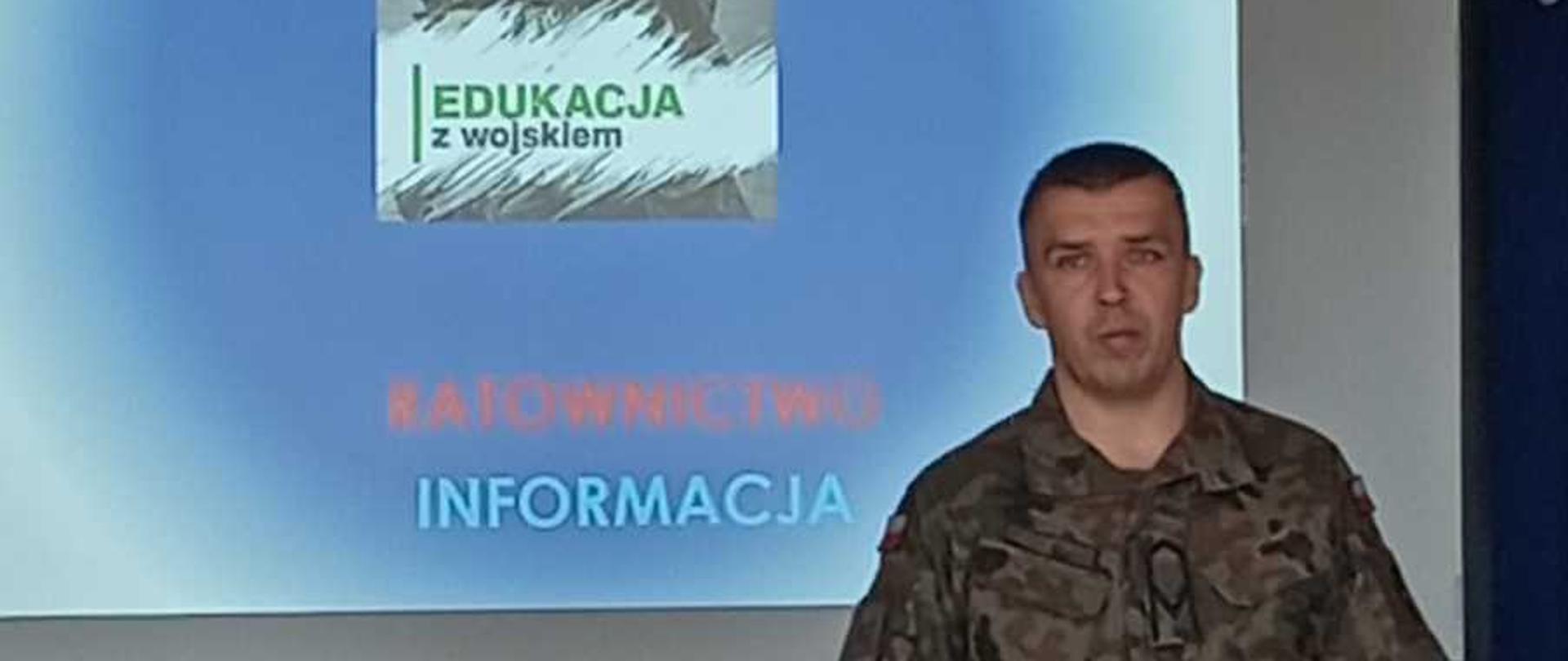 Zdjęcie przedstawia prelekcję prowadzoną przez żołnierza w sali lekcyjnej lub konferencyjnej.Główne elementyPrelegent: W prawej części kadru stoi mężczyzna w średnim wieku, ubrany w polski mundur wojskowy w kamuflażu pantera. Jest zwrócony przodem do słuchaczy.Ekran projekcyjny: Za żołnierzem znajduje się duży, jasny ekran, na którym wyświetlana jest prezentacja multimedialna. Nad ekranem widać podwieszony pod sufitem projektor.Publiczność: Na pierwszym planie, tyłem do obiektywu, widoczne są głowy i ramiona siedzących w ławkach słuchaczy (młodzieży).Treść prezentacji na ekranieNa slajdzie widnieją napisy w języku polskim, ułożone pionowo:BEZPIECZEŃSTWO (duży, niebieski napis na samej górze)ALARMOWANIE (zielony napis)OCHRONA (pomarańczowy napis)W centrum znajduje się grafika z logo programu: „EDUKACJA z wojskiem”RATOWNICTWO (czerwony, słabo widoczny napis)INFORMACJA (jasnoniebieski napis na dole)Kadr jest statyczny, a światło w pomieszczeniu jest skupione głównie na ekranie prezentacji.