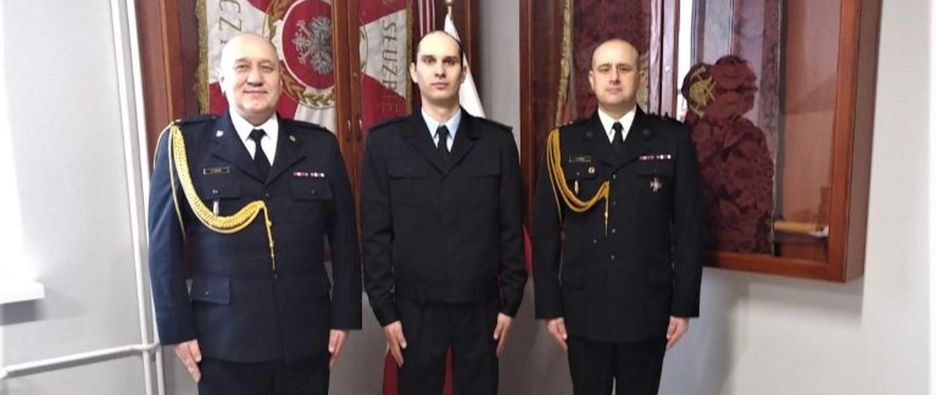 Zdjęcie przedstawia komendanta, zastępcę w umundurowaniu galowym oraz strażaka w umundurowaniu służbowym, który złożył ślubowanie. W tle witryny ze sztandarem komendy. 