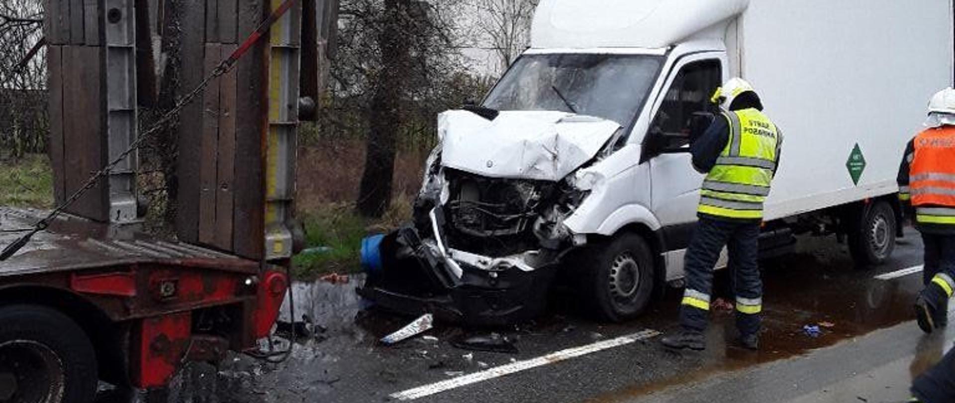 
Na zdjęciu widoczny biały samochód dostawczy z uszkodzoną komorą silnika. Samochód ten najechał na tył ciągnika siodłowego. Przy biały samochodzie stoi strażak w kamizelce odblaskowej.
