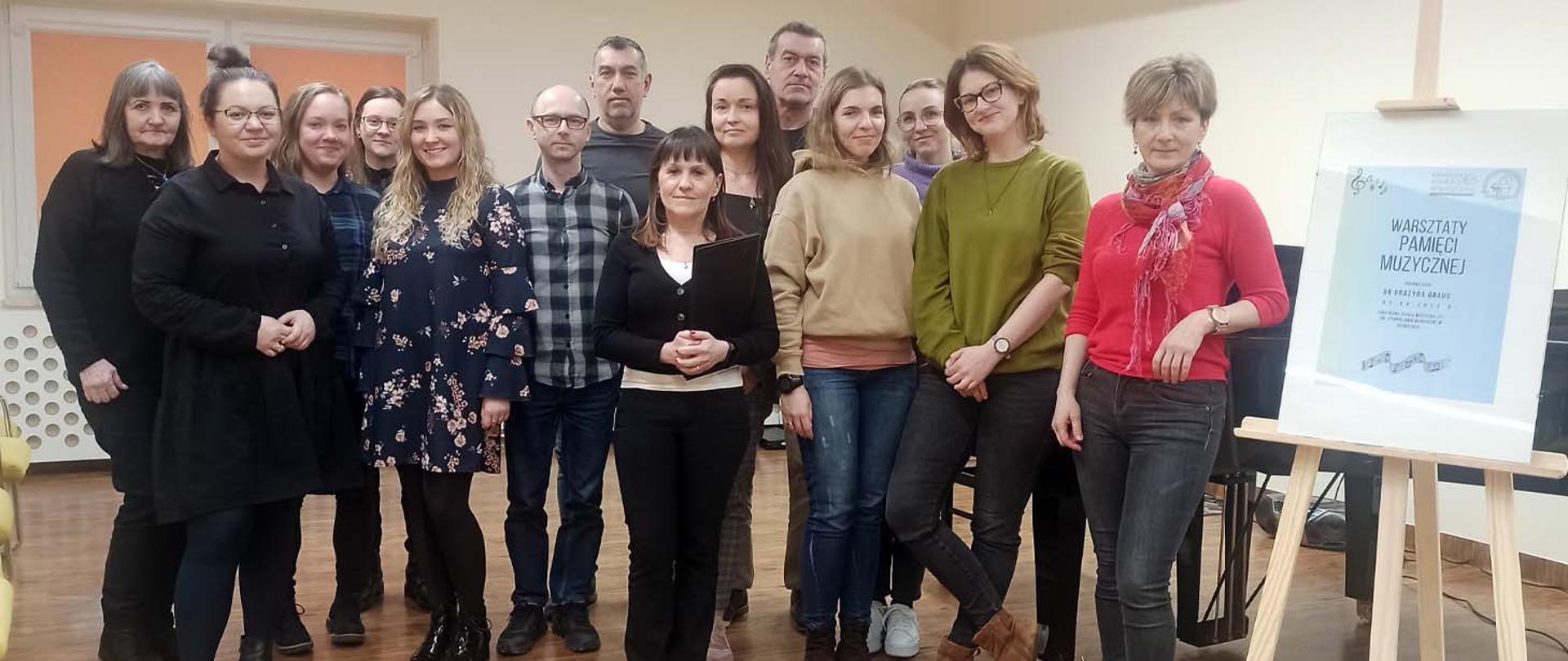 Uczestnicy warsztatów stoją w auli szkoły, po prawej stronie plakat z logotypami organizatorów.