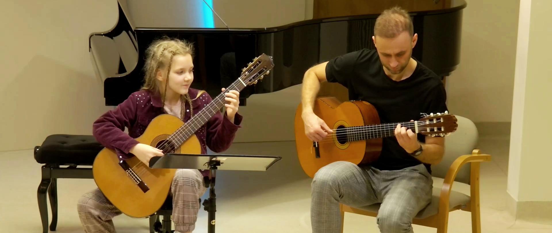 Zdjęcie. Na pierwszym planie z prawej strony młoda gitarzystka gra na gitarze. Przed nią stoi pulpit z nutami. Towarzyszy jej - z lewej strony - nauczyciel, który również gra na gitarze. W tle stoi czarny fortepian. Koncert odbywa się w Sali Kameralnej PSM w Nysie.