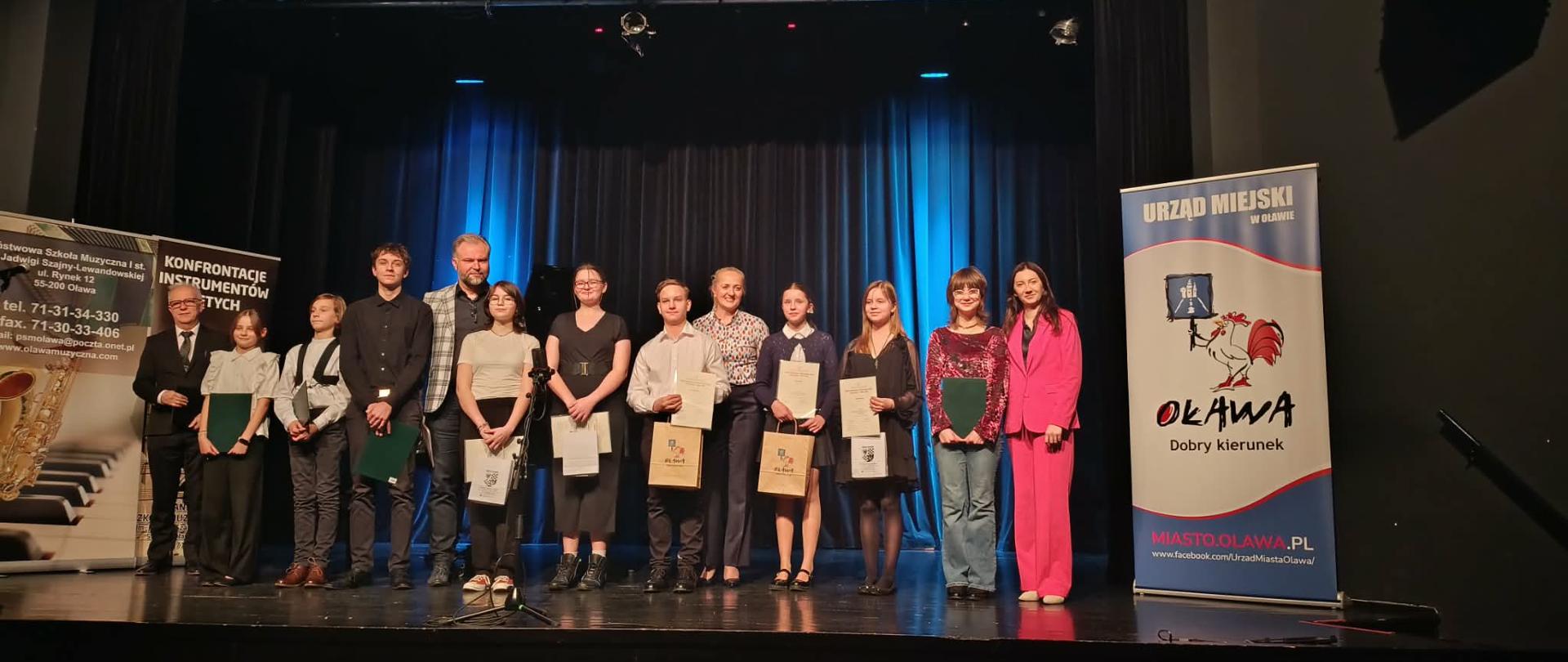 Uczniowie stoją na scenie kina z dyplomami, po bokach banery reklamowe