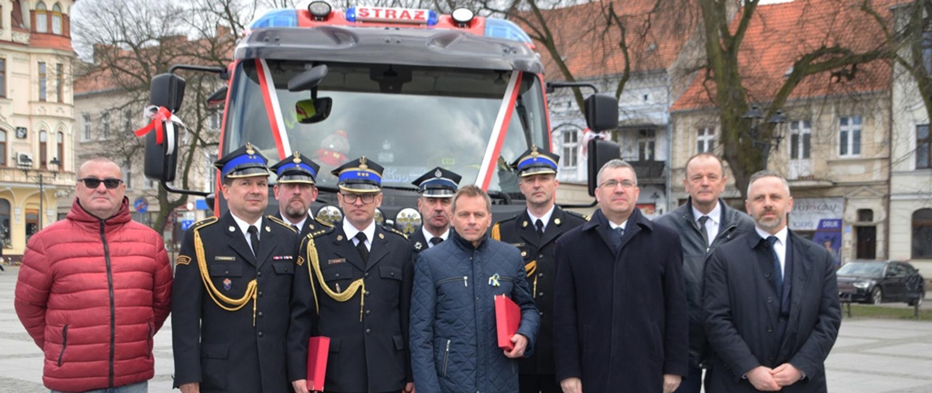 Na zdjęciu na płycie Chełmińskiego Rynku znajdują się zaproszeni goście podczas uroczystego apelu z okazji przekazania i poświęcenia pojazdu dla OSP Chełmno. Zaproszeni goście stoją na tle przekazywanego pojazdu podczas pamiątkowego zdjęcia.