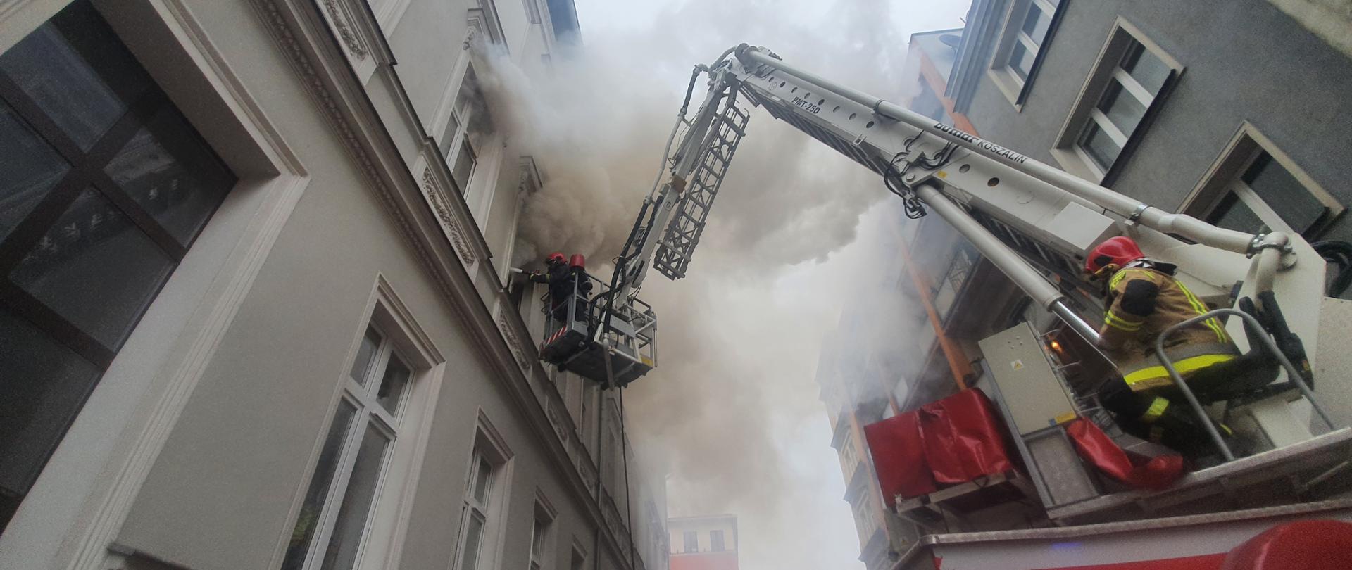 Rozstawiona drabina mechaniczna straży pożarnej podczas gaszenia pożaru kamienicy. Na pierwszym palnie operator drabiny strażak, w perspektywie na wysokości w koszu dwoje strażaków gasi pożar przez okno mieszkania. Wokoło panuje duże zadymienie. 