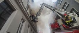 Rozstawiona drabina mechaniczna straży pożarnej podczas gaszenia pożaru kamienicy. Na pierwszym palnie operator drabiny strażak, w perspektywie na wysokości w koszu dwoje strażaków gasi pożar przez okno mieszkania. Wokoło panuje duże zadymienie. 