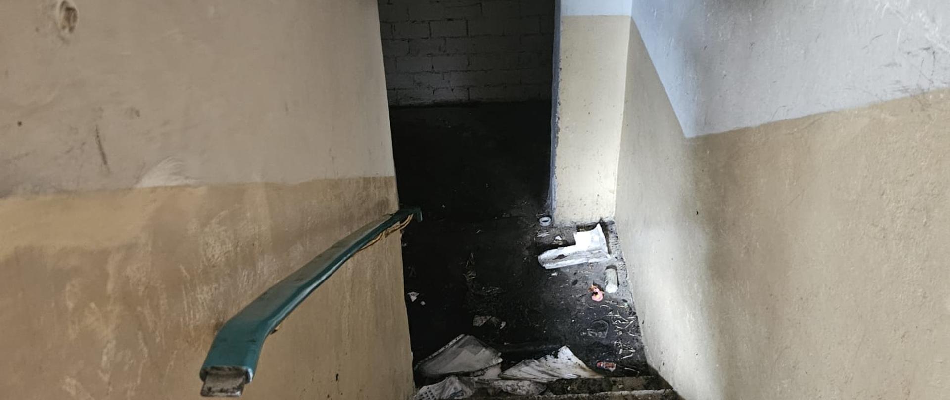 Wąska klatka schodowa prowadząca w dół do ciemnego pomieszczenia. Ściany i sufit są silnie okopcone czarną sadzą, szczególnie po prawej stronie i na suficie. Na betonowych schodach leżą śmieci oraz pozostałości po pożarze. Po lewej stronie znajduje się niebieska poręcz. Na dole widoczna jest ciemna podłoga i ceglana ściana w tle. Zdjęcie przedstawia zniszczenia i zabrudzenia po pożarze lub silnym zadymieniu