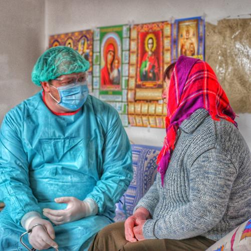 Lekarz w stroju ochronnym, maseczce i goglach rozmawia z pacjentką w kolorowej chuście w domu, w którym widnieją obrazy świętych.