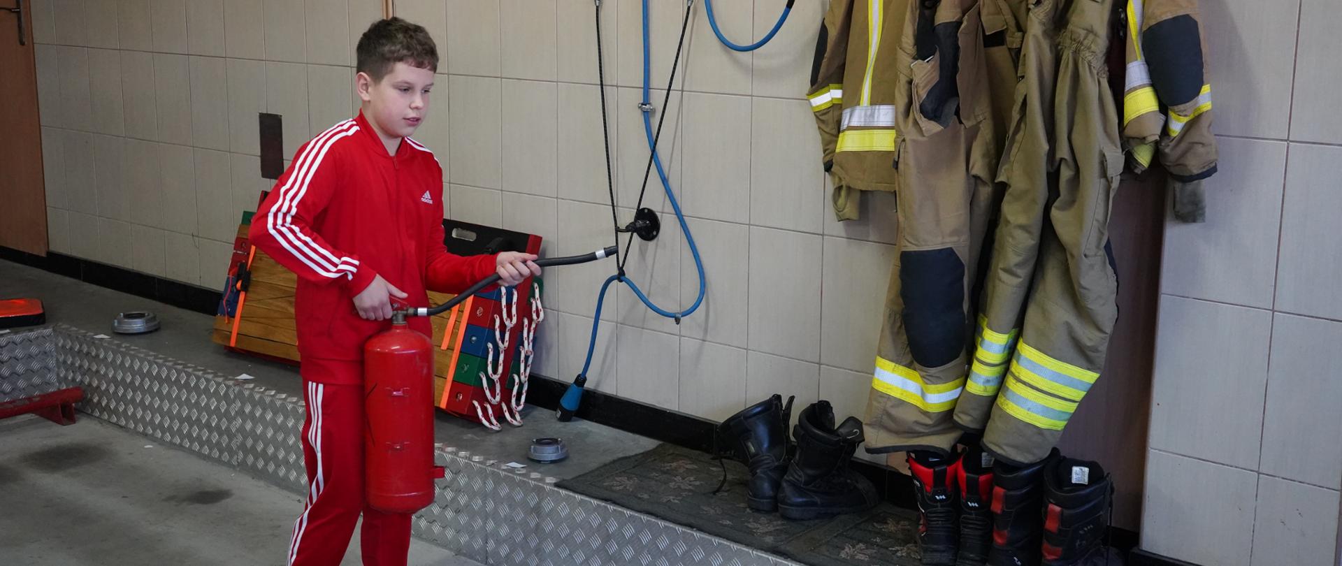 Remiza OSP. Jeden z uczestników eliminacji trzyma w rękach gaśnicę, przy pomocy której symuluje gaszenie pożaru. Chłopiec ma na sobie czerwony dres z białymi pasami na nogawkach i rękawach. Na wieszaku umiesczonym na ścianie wiszą ubrania specjalne. Pod nimi stoją 3 pary butów. 