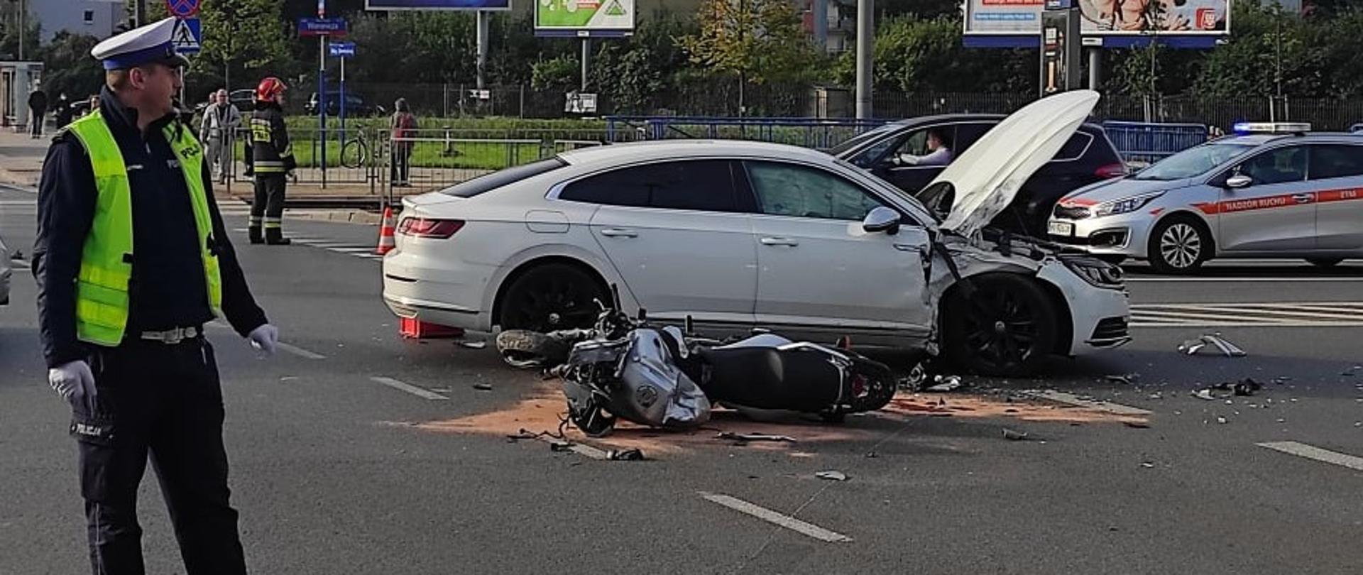 Na zdjęciu widoczny jest motocykl oraz samochód osobowy, pojazdy brały udział w wypadku oraz policjant.