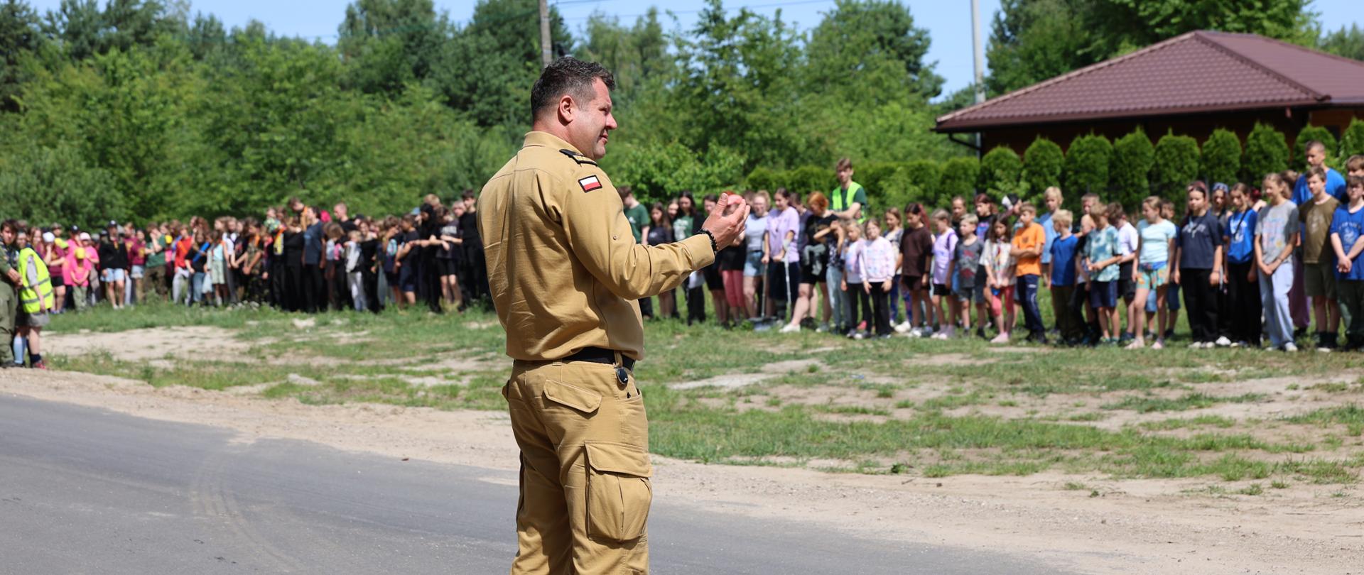 Na zdjęciu komendant PSP w piaskowym mundurze stoi na drodze i zwraca się do uczestników ewakuacji. Po drugiej stronie drogi zgromadzeni są harcerze oraz kadra obozu. W tle znajduje się altana oraz las. Pogoda jest słoneczna.