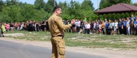 Na zdjęciu komendant PSP w piaskowym mundurze stoi na drodze i zwraca się do uczestników ewakuacji. Po drugiej stronie drogi zgromadzeni są harcerze oraz kadra obozu. W tle znajduje się altana oraz las. Pogoda jest słoneczna.