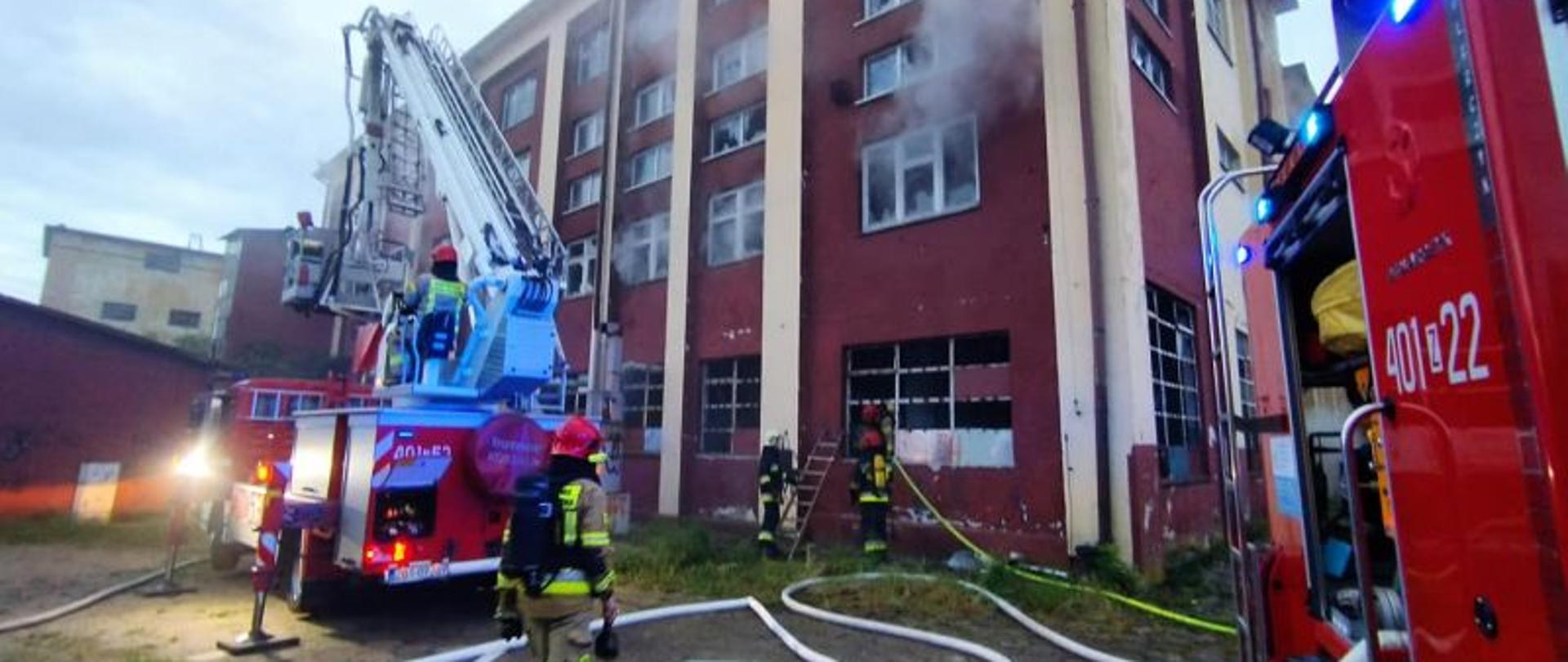 Widok na miejsce akcji. Z lewej rozstawiony podnośnik a z prawej widoczny kawałek wozu gaśniczego. Na środku strażacy a w tle wysoki opuszczony budynek zakładu. Budynek ma dużo okien z których wydostaje się dym.
