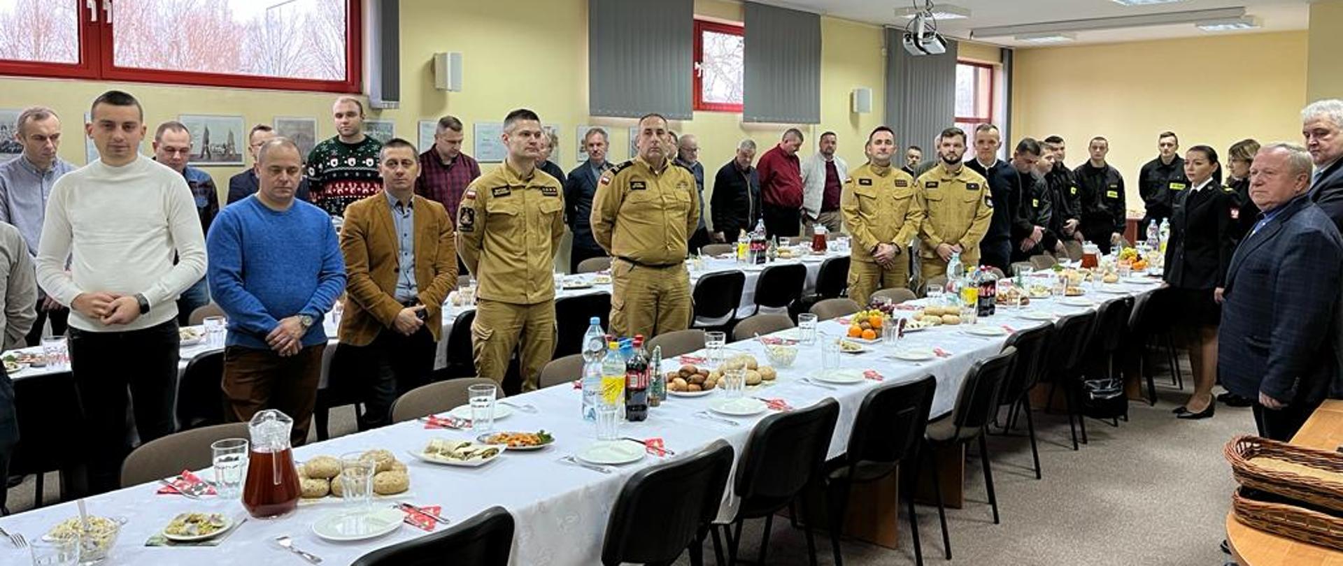 Strażacy, pracownicy cywilni i emeryci KP PSP w Grodzisku Maz. oraz zaproszeni goście przy zastawionych stołach w trakcie spotkania wigilijnego słuchają przemów komendanta i zaproszonych gości. 