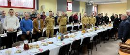 Strażacy, pracownicy cywilni i emeryci KP PSP w Grodzisku Maz. oraz zaproszeni goście przy zastawionych stołach w trakcie spotkania wigilijnego słuchają przemów komendanta i zaproszonych gości. 