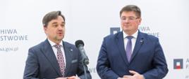 Pod ścianką z napisem Archiwa Państwowe stoi wiceminister Rzymkowski i mężczyzna w garniturze, który mówi do mikrofonu na stojaku.