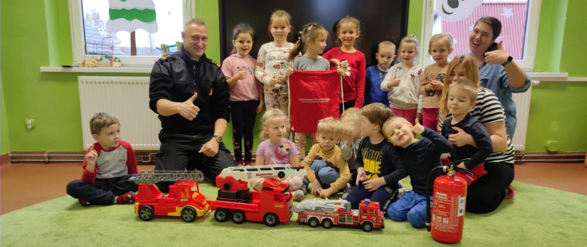 Na zdjęciu znajduje się strażak w ubraniu służbowym oraz dzieci w pomieszczeniu