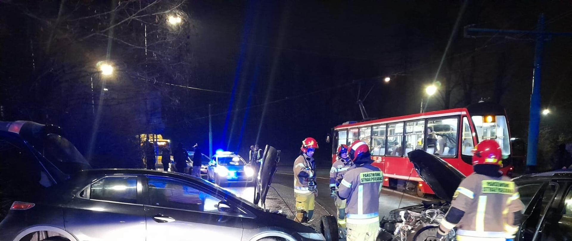 Na zdjęciu na pierwszym planie znajdują się dwa uszkodzone samochody osobowe skierowane do Siebie oraz strażacy wykonujący swoje czynności, za nimi w w tle tramwaj radiowóz Policji.