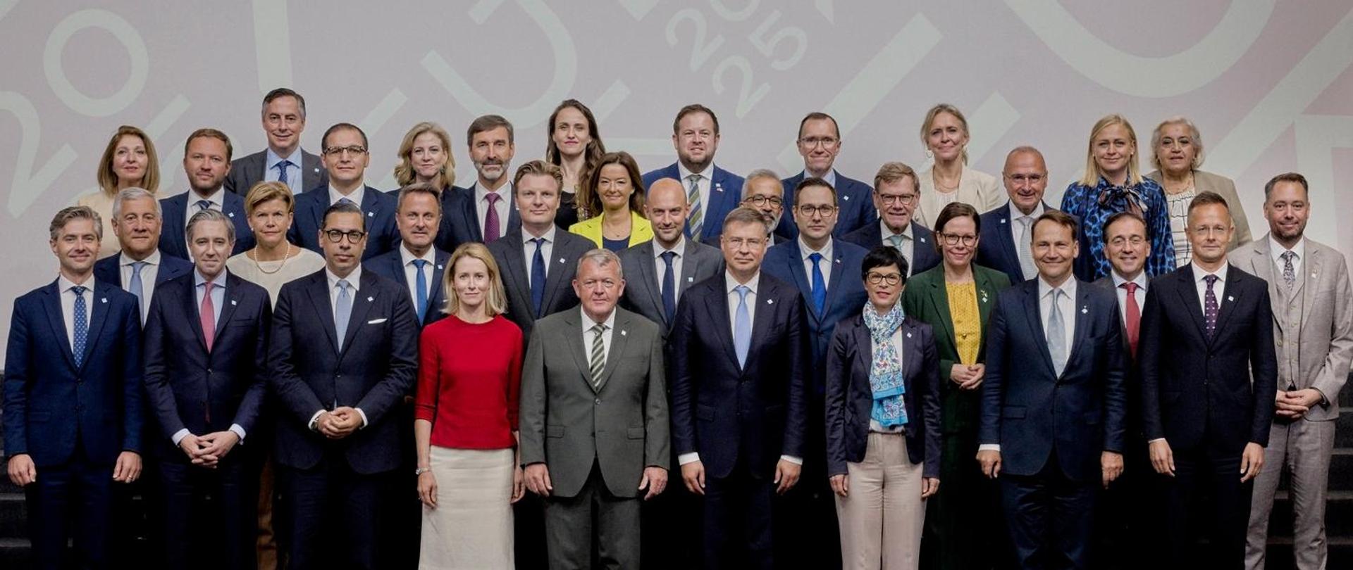 Zdjęcie grupowe przedstawia szefów dyplomacji państw członkowskich Unii Europejskiej, osoby ubrane w stroje formalne stoją na schodach w czterech rzędach na tle baneru z logotypem duńskiej prezydencji w Radzie Unii Europejskiej