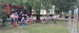 Dzieci z prądownicami i hydronetkami podają wodę do 2 makiet domów. Za nimi samochód gaśniczy. 
