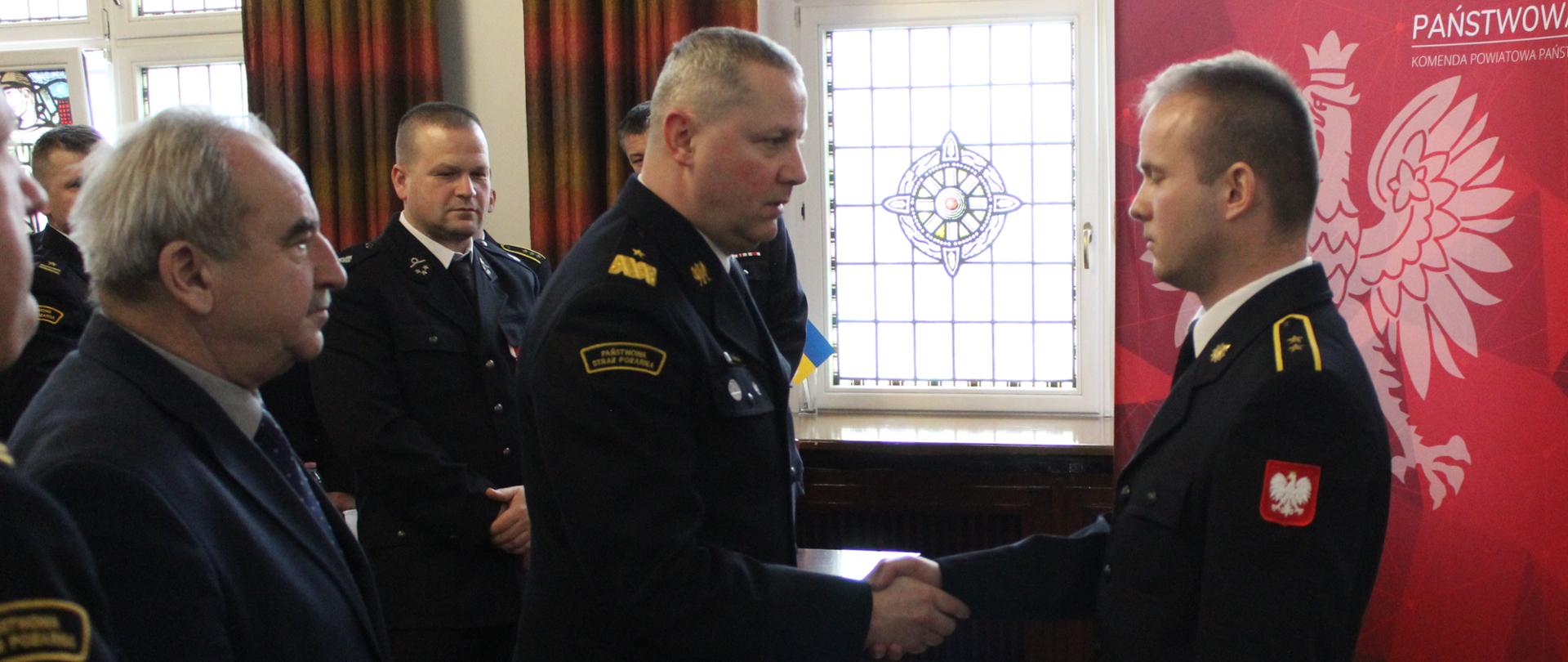 strażak gratuluje drugiemu strażakowi, ściska mu dłoń w tle grupa mężczyn i orzeł na czerwonym tle 