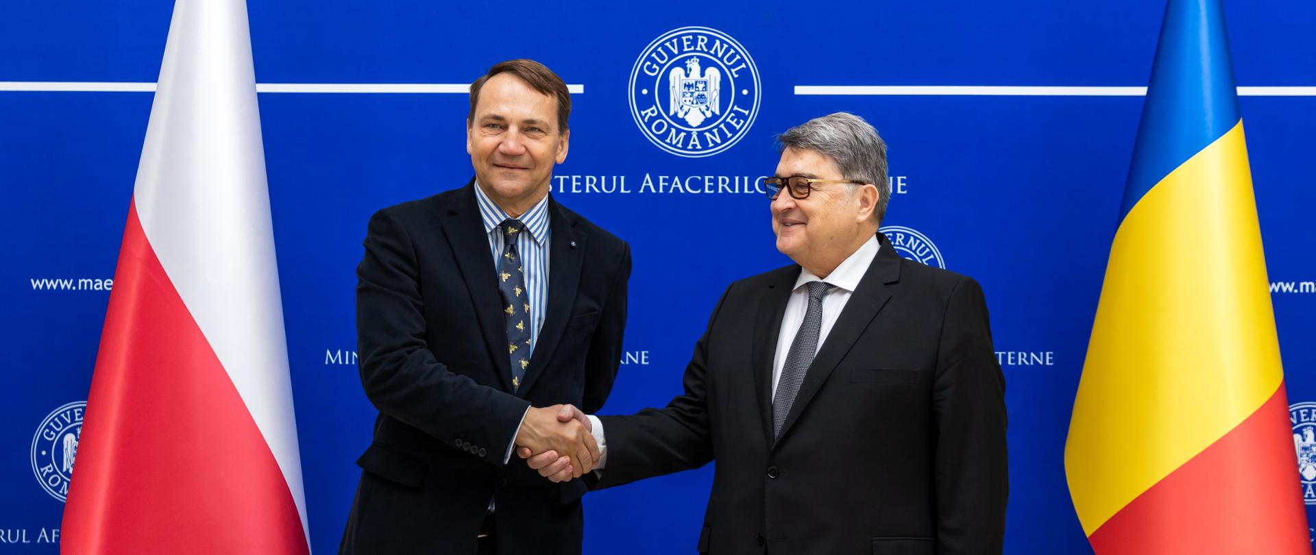 Dwóch mężczyzn, szefowie dyplomacji Polski i Rumunii, stoją na tle ścianki prasowej i flag Polski i Rumunii oraz podają sobie ręce.