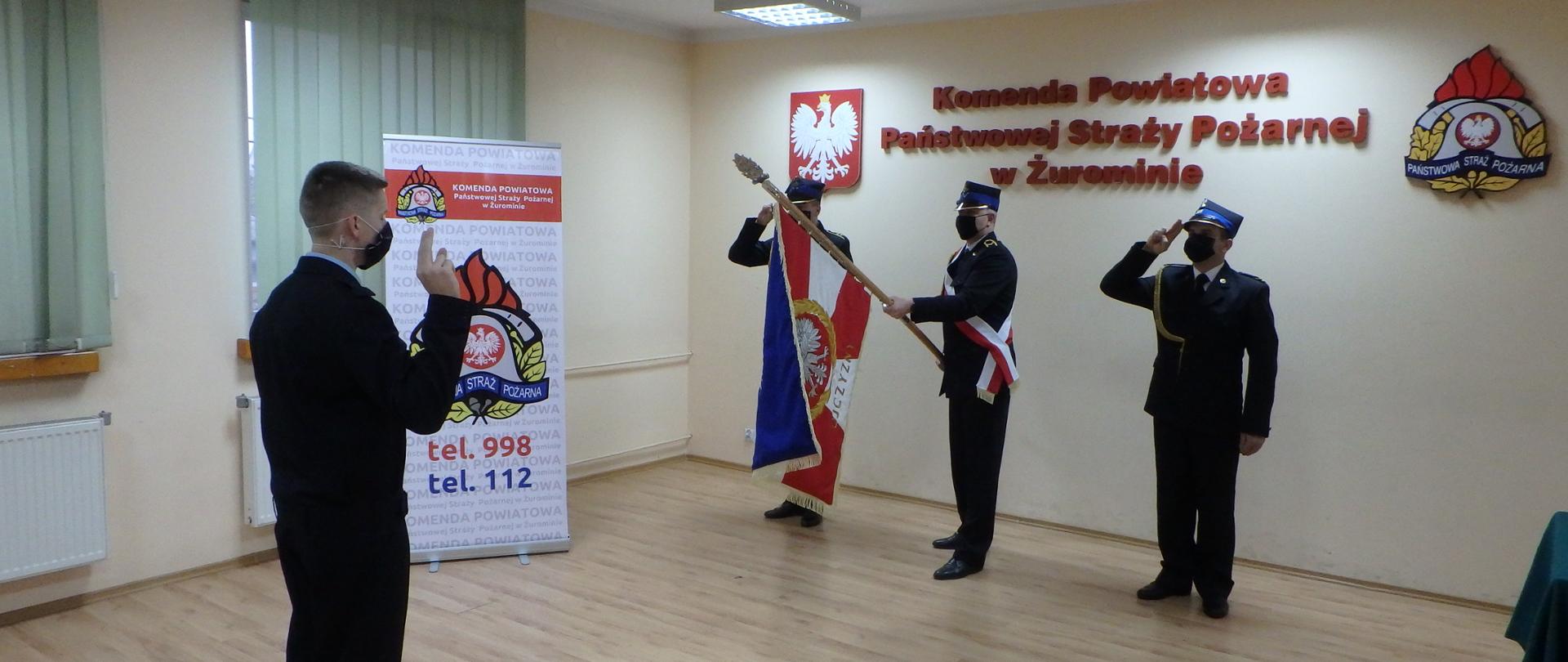 W centralnej części zdjęcia znajduje się strażak składający ślubowanie z podniesioną ręką i palcami złożonymi jak do salutowania. W dalszej części zdjęcia naprzeciw strażaka składającego ślubowanie jest ustawiony poczet sztandarowy składający się z trzech strażaków. Po środku pocztu stoi strażak trzymający pochylony pod kątem 45 stopni sztandar Komendy Powiatowej Państwowej Straży Pożarnej w Żurominie, strażacy stojący po obu stronach strażaka ze sztandarem stoją salutujący strażacy. Na ścianie za pocztem widnieje napis na ścianie Komenda Powiatowa Państwowej Straży Pożarnej w Żurominie, z lewej strony napisu jest umieszczone godło Rzeczypospolitej Polskiej a z prawej strony napisu umieszczone jest logo Państwowej Straży Pożarnej.