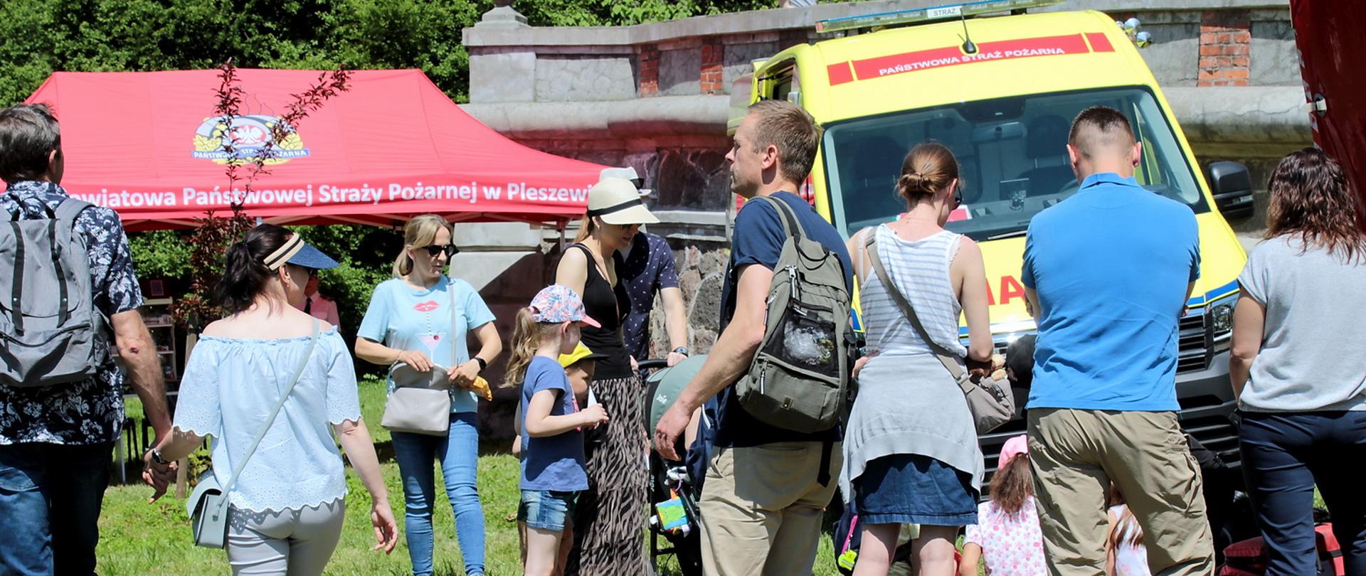 Grupa dzieci i dorosłych stojące przed stoiskiem edukacyjnym PSP, widoczny czerwony namiot stoiska z napisem Komenda Powiatowa Państwowej Straży Pożarnej w Pleszewie oraz żółta karetka medyczna