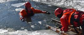 Na zdjęciu widoczni dwaj strażacy na skutym lodzie akwenie wodnym. Obaj w suchych skafandrach koloru pomarańczowego w czerwonych kaskach ochronnych. Jeden ratownik z użyciem drabin nasadkowych ewakuuje z wody drugiego strażaka.