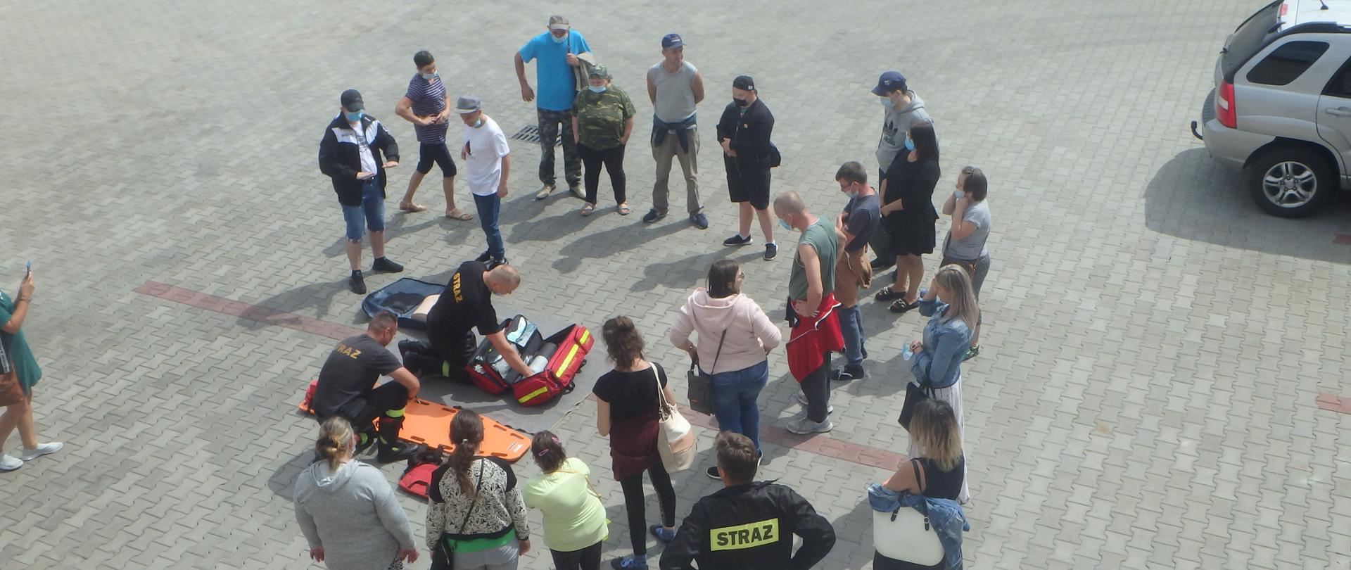 Dwóch strażaków klęczy przed deską ortopedyczną i torbą medyczną na środku placu przed komendą. Wokół nich stoją ludzie. Wokoło widać też zaparkowane samochody.