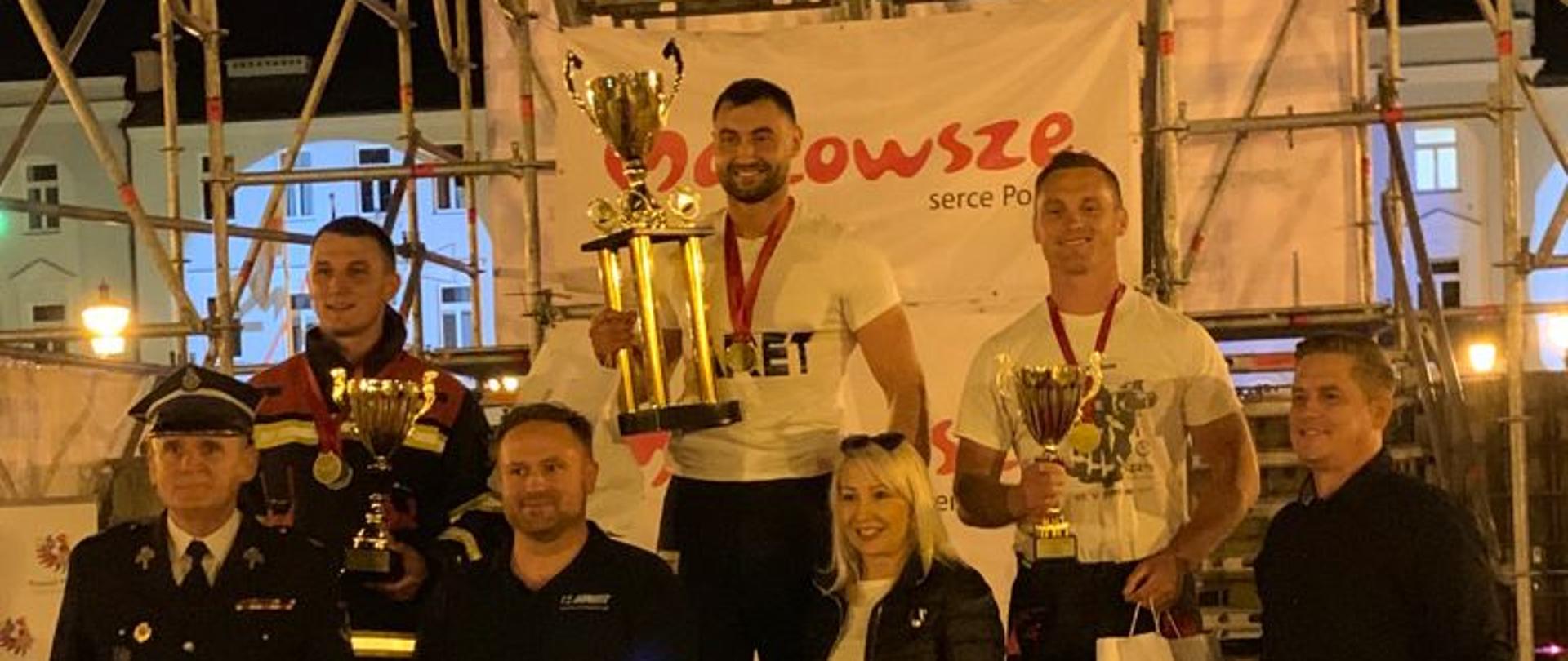 Trzech strażaków stojących na podium zawodów po dekoracji. Cztery osoby stoją przed podium. W tle wieża sportowa z zawieszonymi na niej banerami sponsorów.