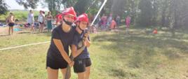 Obóz szkoleniowo-wypoczynkowy MDP w Brennie. Na plaży odbywają się zawody sportowo-pożarnicze. Na pierwszym planie dwie obozowiczki w czerwonych hełmach, które podają wodę z węża i prądownicy. W tle inni obozowicze oraz drzewa.