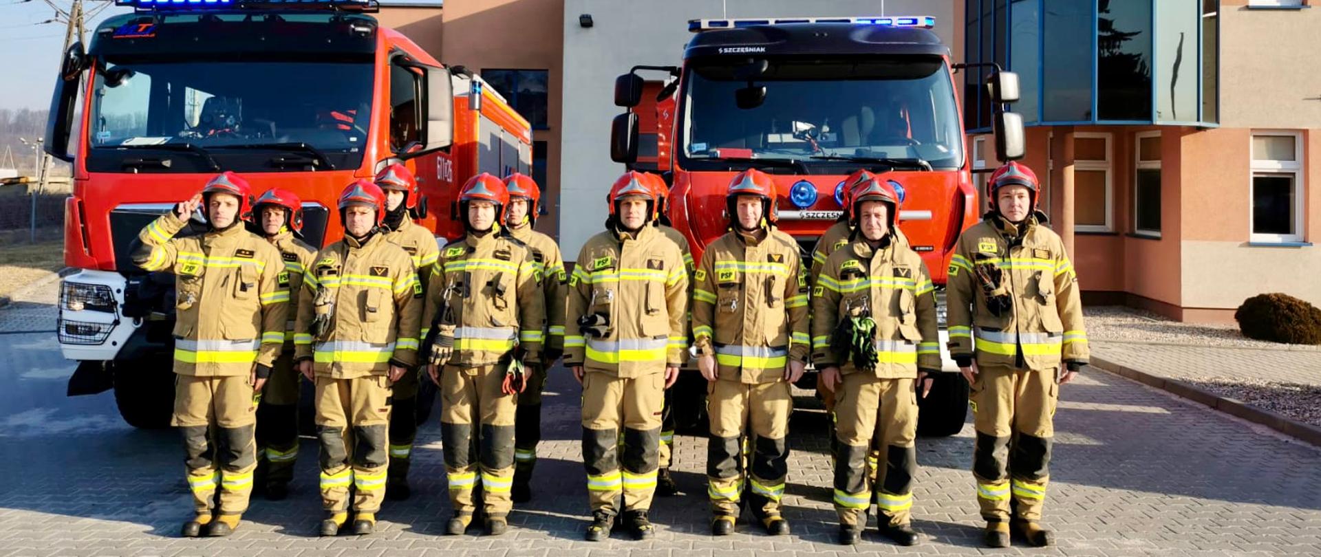 umundurowani strażacy na tle pojazdów gaśniczych oddający honory poprzez salutowanie