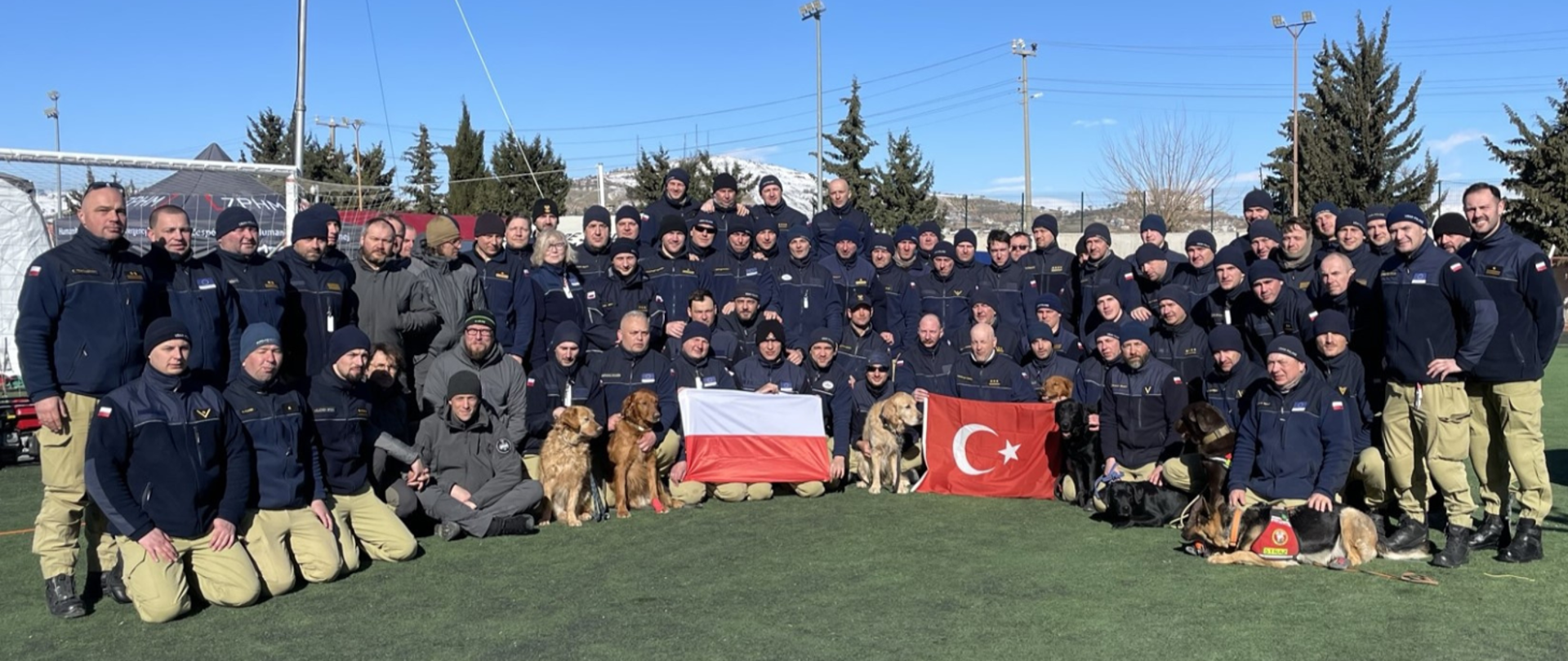 Grupowe zdjęcie polskich strażaków wraz z psami ratowniczymi. w rękach trzymają flagi Polski i Turcji
