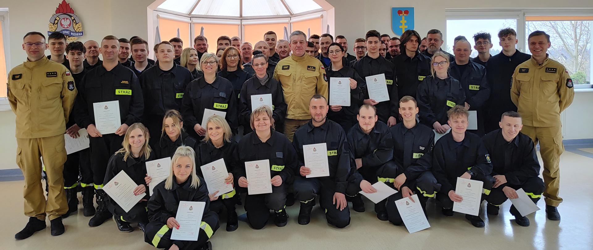 Zdjęcie grupowe strażaków OSP oraz komendanta PSP, dowódcy JRG i zastępcy na tle godła Polski i logo PSP, którzy pozytywnie skończyli kurs OSP. 