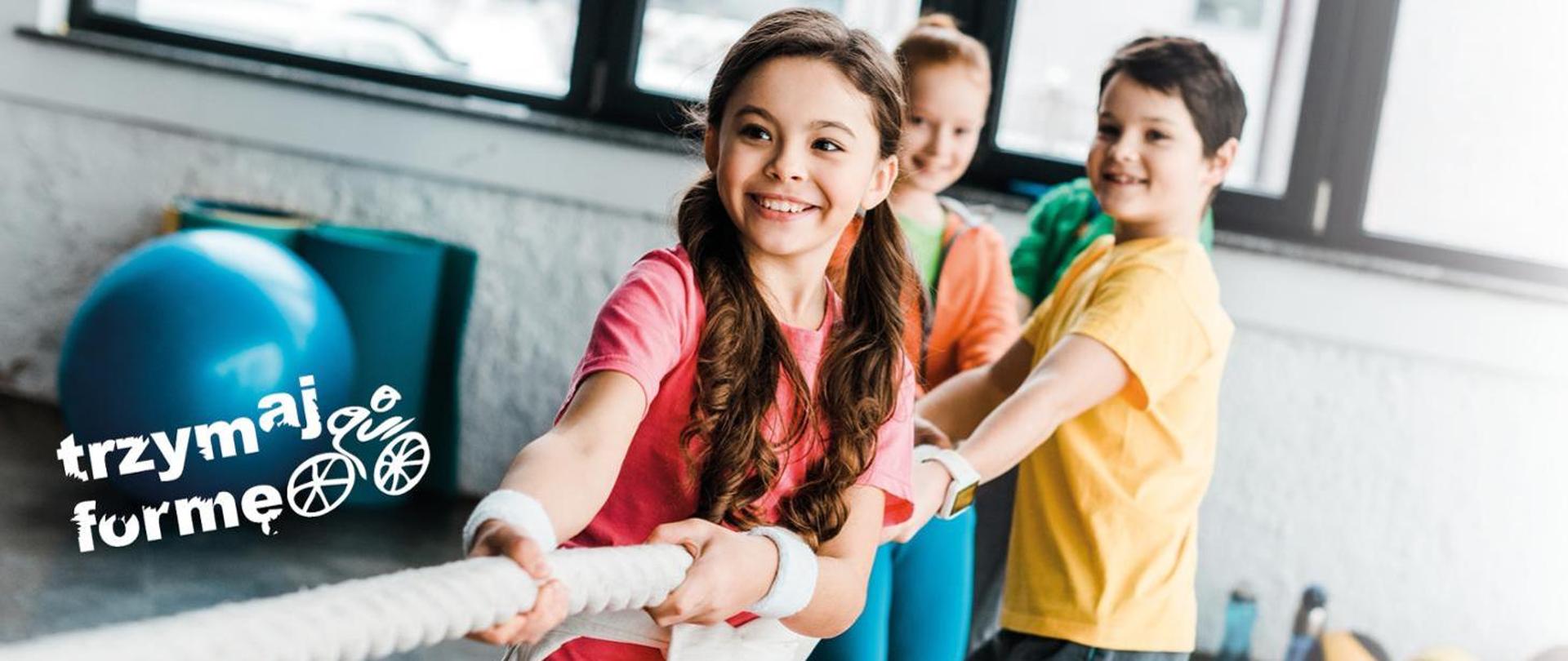  3 dzieci przeciągających linę na sali gimnastycznej z boku napis Trzymaj Formę