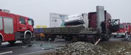 Samochód ciężarowy uderzył w naczepę drugiego samochodu ciężarowego stojącą w poprzek drogi. Samochody pożarnicze stojące po obu stronach zdarzenia.