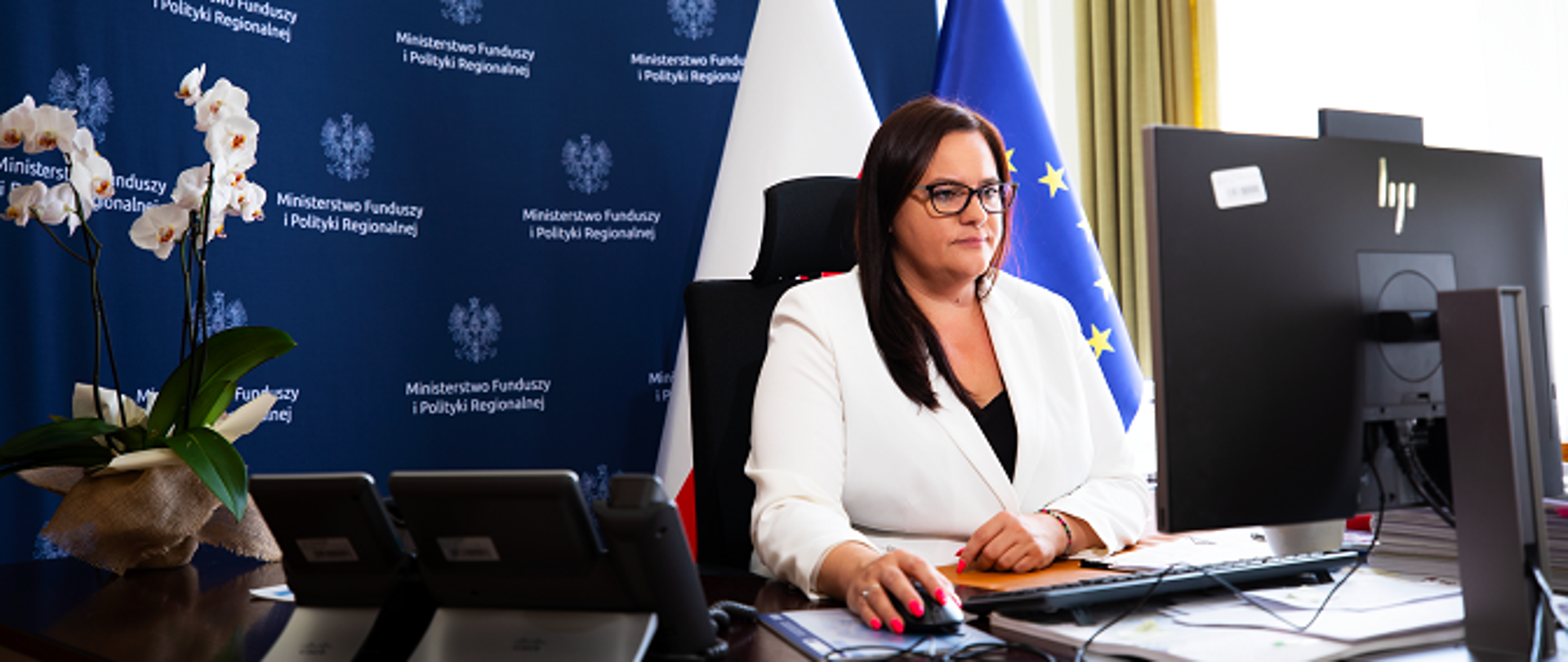Wiceminister Małgorzata Jarosińska-Jedynak siedzi za biurkiem, przed komputerem. Za plecami ścianka z logo MFiPR