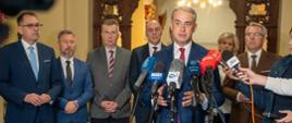Na zdjęciu wiele osób. W środku wysoki mężczyzna w garniturze. Przed nim stoją mikrofony. Trwa konferencja prasowa.