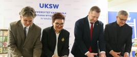 Minister Czarnek, dwóch mężczyzn i kobieta ubrana na czarno razem przecinają nożyczkami wstęgę rozciągniętą miedzy dwoma mosiężnymi słupkami.