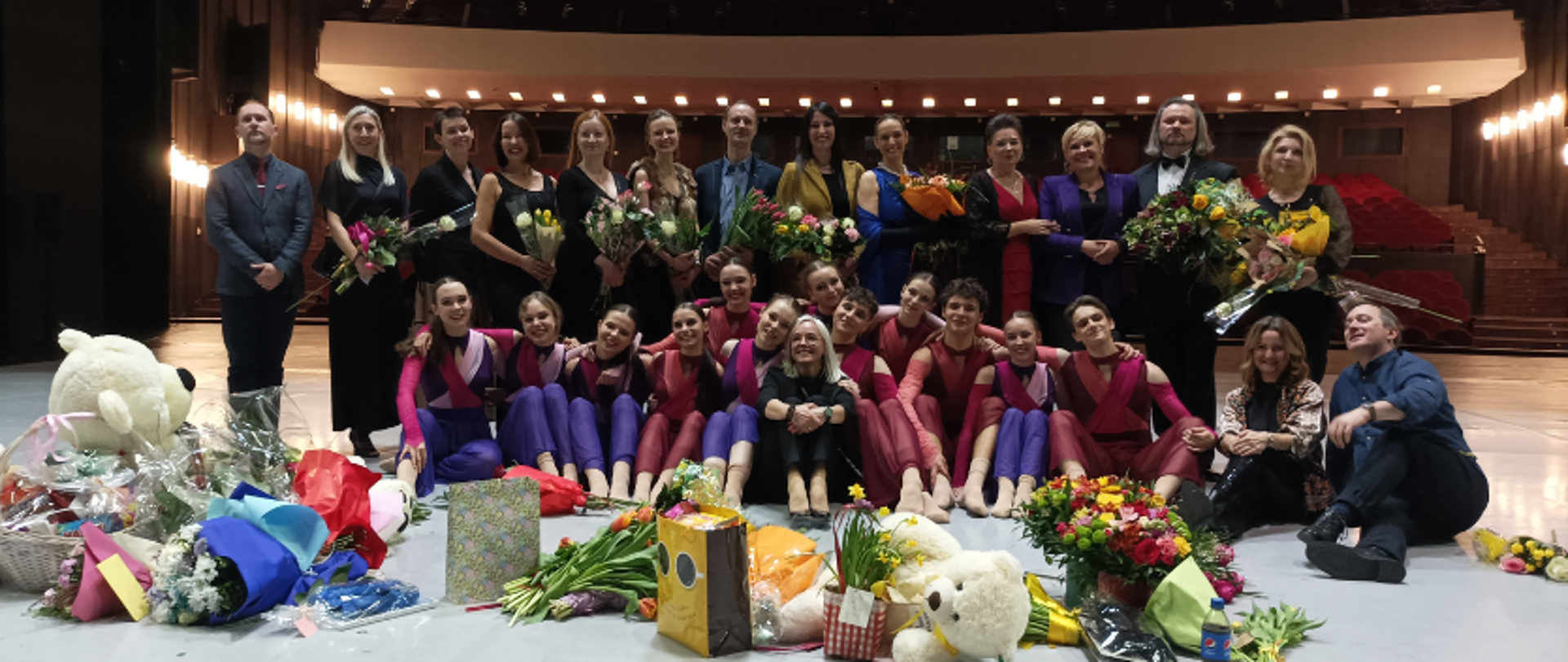 zdjęcie: uczniowie 9 klasy baletowej szkoły baletowej w Łodzi oraz ich pedagodzy na scenie Teatru Wielkiego w Łodzi po zakońzzeniu XVII Koncertu Galowego szkoły. Uczniowie w kostiumach scenicznych siedzą na scenie, za nimi stoją pedagodzy tańca. Na scenie przed nimi lezą również kwiaty i prezenty.