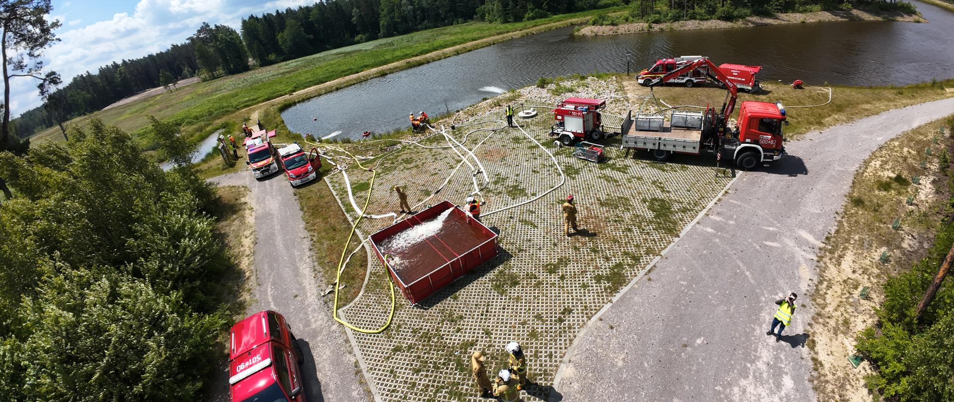 Panoramiczne zdjęcie lotnicze przedstawiające główny punkt ćwiczeń zlokalizowany na cyplu obok dużego zbiornika wodnego. Na placu z geokraty rozstawiony jest duży, prostokątny, czerwony przenośny zbiornik buforowy, do którego z rzeki pompowana jest woda (widoczne białe spienione strumienie). Wokół zbiornika rozłożona jest skomplikowana sieć białych węży strażackich. Na placu stoi kilka pojazdów: czerwony pickup, wóz strażacki z dźwigiem HDS, mniejsza autolaweta oraz inne samochody ratownicze. Wokół sprzętu operuje kilkunastu strażaków w strojach bojowych oraz odblaskowych kamizelkach.