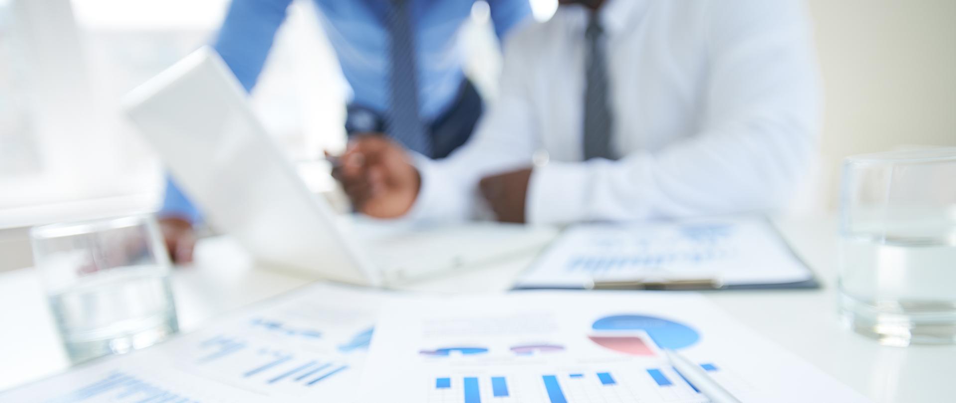 Dokumenty z wykresem i piórem na tle dwóch pracowników.