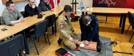 Na zdjęciu na pierwszym planie przy manekinie do pierwszej pomocy znajdują się dwie osoby, jedna to strażak, ćwiczą resuscytację krążeniowo-oddechową. Na drugim planie siedząc przy stoliku 3 osoby obserwują ćwiczenia.